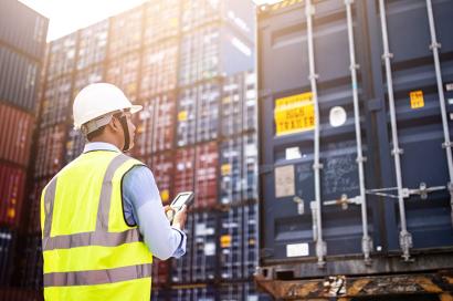 A worker in a safety vest and helmet is using a smartphone while inspecting shipping containers at a port.
